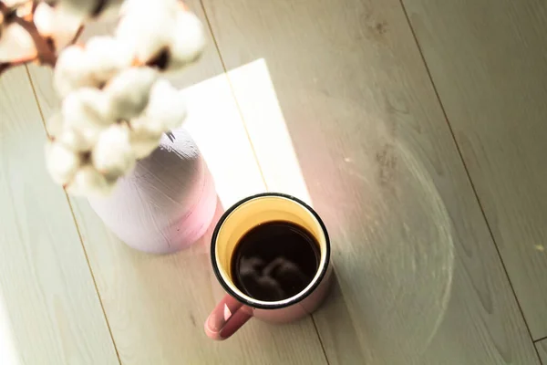A cup of black coffee in a ceramic cup, mug in focus, a branch of white cotton flower in a vase in defocus on a white parquet floor background. Cozy home at autumn season. Sunlight. Place for text.