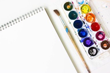 Açılan eskiz defteri, beyaz suluboya kağıtlar, çok renkli boyalar ve masanın üstünde duran fırçalar. Resim dersi. Eğitim sanatı. Okula dönelim. Düz çalışmak için aletler. Metin için yer.