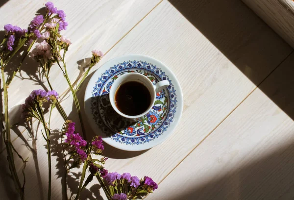 Turkish coffee in traditional blue porcelain cup at saucer on a white wooden floor top view. Contrasting shadows. Hot espresso drink in a morning light flatly. A composition of dried purple flowers.