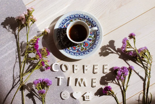 A cup of Turkish coffee on a white table with a silver tablecloth. Coffee time text inscription, smiley face, heart, home symbols top view. Dried purple lilac flowers flat lay. Beautiful composition.