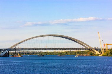 Kyiv, Ukrayna. 2 Mayıs 2021. Dnipro nehrinde mavi bir suyun üzerine büyük kemerli bir köprü inşa etmek. İnşaat vinci, yaz günü mavi gökyüzü arka planına karşı bir ufukta. Modern şehir mimarisi. 