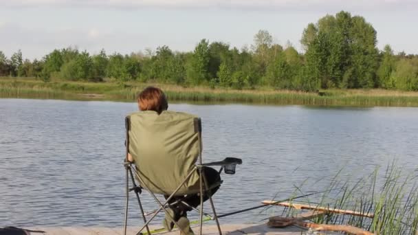 Fischerin beim Fischfang. Eine Frau sitzt mit einer Angelrute in der Hand auf einem klappbaren Touristenstuhl am Flussufer und fängt Fische. Holzsteg, grünes Schilf an einer Böschung. Wasser fließt. Freizeitwochenende. — Stockvideo
