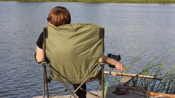 Μια γυναίκα ψαρεύει με καλάμι σ 'ένα καλοκαίρι στην όχθη του ποταμού. Γυναίκα χαλαρώνει σε μια πτυσσόμενη καρέκλα κάμπινγκ, βλέποντας μια ροή του νερού. Όμορφο τοπίο. Ηρεμία, αρμονία, αθλητισμός έννοια σε εξωτερικούς χώρους. — Αρχείο Βίντεο