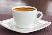 Kulatý bílý keramický šálek s kávovou hnědou pěnou cappuccino latte na čtvercovém podšálku na stole. Teplý aromatický nápoj v kavárně, restaurace ráno, oběd na podzim. Motiv nápojů.