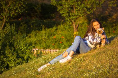 Gümüş renkli yağmurluk giyen, uzun saçlı, güzel bir kız güneşli bir günde parkın çimlerinde uzanmış, küçük bir Yorkshire teriyerine sarılıyor, komik köpek yavrusu. İnsanlar ve hayvanlar, hayvanları severler.