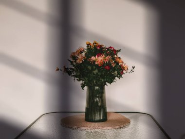 Minimalist bir çiçek buketi gibi yeşil cam bir vazoda dokuma bir paspasın üzerinde duruyor. Pencere gölgeleri modern estetik bir iç tasarım yaratıyor. Konforlu ev.