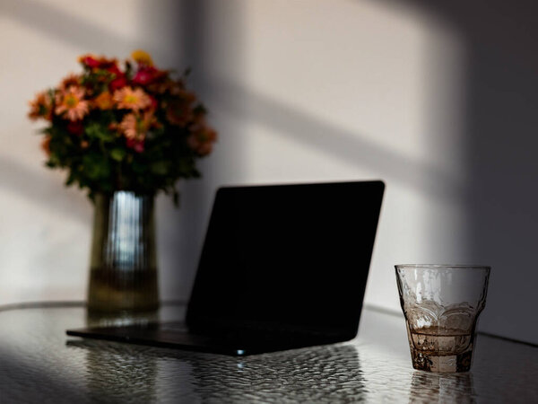 Уютное домашнее рабочее место с ноутбуком, стаканом воды и вазой цветов в теплом естественном свете. Минималистичные настройки удаленной работы, мягкие тени, спокойная атмосфера идеально подходит для продуктивной работы на дому.