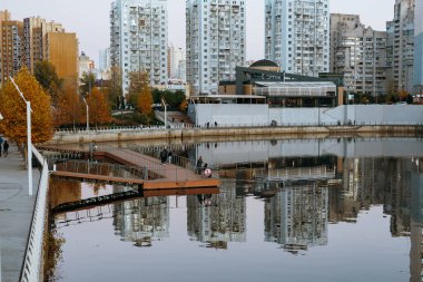 Kyiv, Ukrayna 'nın 25 Ekim 2025 Cityscape' inde su ve modern mimaride yansımalar var. Sakin su binaları yansıtıyor ve güzel bir şehir manzarası yaratıyor. İnsanlar iskelede yürüyor..