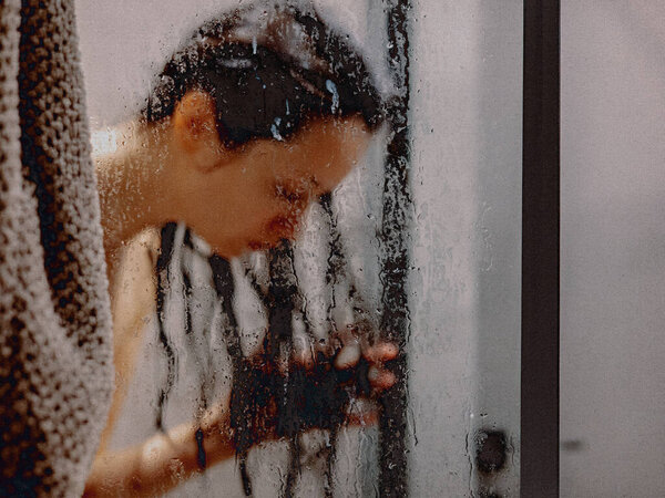 Woman in shower behind glass with water droplets, spa and wellness concept. A white woman is seen behind a glass shower door, creating a sense of privacy and relaxation. Dark hair care Beauty products