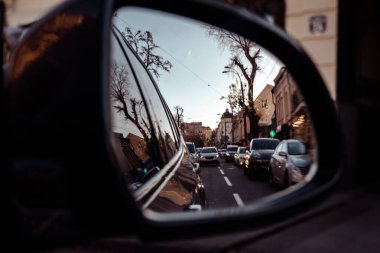 Trafik ve klasik mimari ile dolu kalabalık bir şehir caddesinin yansıması araba yan aynasında, gidip gelişi, yolculuğu ve şehir hayatını sembolize ediyor. Kentsel seyahat perspektifi. Dikiz aynası. Otomobil gezisi.