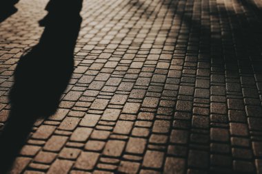 İnsanların siluetlerinin ve kaldırımda yürüyen uzun gölgelerin yüksek kontrastlı görüntüleri. Dramatik ışıklandırma atmosferik bir etki yaratıyor, günlük şehir yaşamının bir anını yakalıyor..