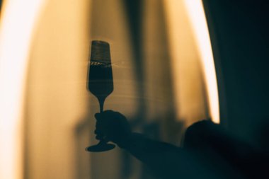 Sıcak bir arka plan duvarına şampanya flütü tutan dramatik bir el silueti. Karanlık trend, estetik, vurgulu ruh hali, kontrast ve özel, gizemli bir an Kutlama içeriği