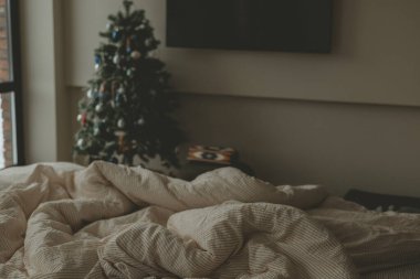 Konforlu bir yatak odası, dağınık bir yatak ve yumuşak bir yatak, sıcak ve rahat bir ev atmosferi yaratıyor. Arka planda süslü bir Noel ağacı olan minimalist bir iç mekan. Kış konforu, kızlık zarı yaşam tarzı.