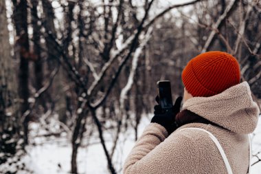 Kırmızı şapkalı ve rahat bej ceketli biri akıllı telefonlu karlı bir ormanın fotoğrafını çekiyor. Bu otantik görüntü, kış yolculuğu ve sosyal medya paylaşımının bir anını yakalıyor. Sosyal medya blogçusu.