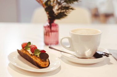 Üzerinde krema ve taze çilek olan bir ekininle birlikte beyaz bir fincan kapuçino ile servis edilen minimalist bir kafe sahnesi. Küçük pembe cam bir vazo rahat ve zarif bir atmosfer yaratır..