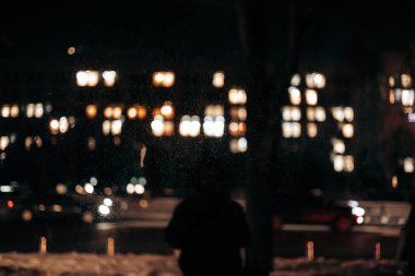 Bulanık gece şehir arka planı, sıcak pencere ışıkları ve karda yalnız bir siluet. Kış kentsel sahnesinde bokeh etkisi, karanlık atmosfer havası, ve gece ya da akşam karanlığında sokak hayatı.