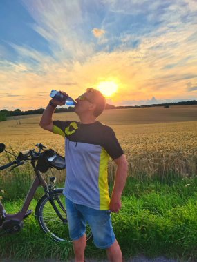 Bisiklet kıyafetleri giymiş, koruyucu başlık takmış, özgür ve mutlu hisseden, şişeden su içen, ılık baharda bisikletin keyfini çıkaran yakışıklı genç bir bisikletçi. Sporcunun yeniden yaratılması.