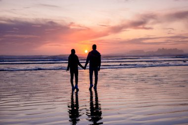Orta yaşlı çift Tofino Vancouver Adası sahilinde gün batımını izliyor. Kanada sahilinde güzel bir gün batımı. Gökyüzünde pembe mor renkler var.