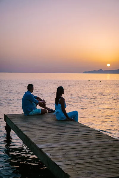 Paar sitzt auf einem Holzsteg und sieht einen bunten Sonnenuntergang am Meer, Männer und Frauen beobachten den Sonnenuntergang auf Kreta Griechenland — Stockfoto