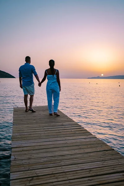 Pareja sentada en un embarcadero de madera, mirando un colorido atardecer en el mar, hombres y mujeres viendo atardecer en Creta Grecia — Foto de Stock