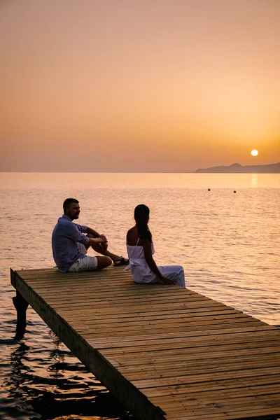 Paar sitzt auf einem Holzsteg und sieht einen bunten Sonnenuntergang am Meer, Männer und Frauen beobachten den Sonnenuntergang auf Kreta Griechenland — Stockfoto