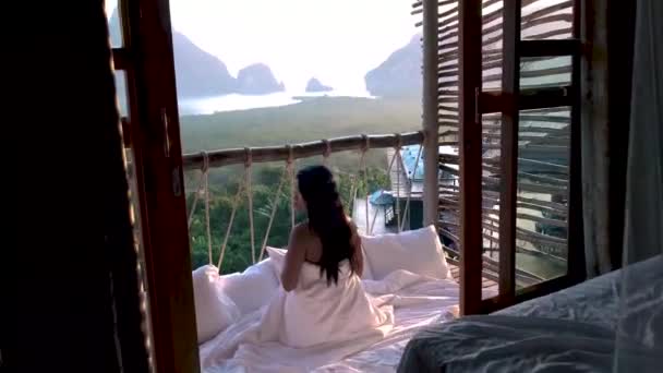 Bahía de Phangnga, mujer asiática despertando en la cama en la selva natural mirando hacia el océano y la selva durante el amanecer en la cabaña de madera en las montañas de Tailandia — Vídeo de stock