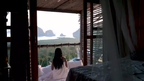 Bahía de Phangnga, mujer asiática despertando en la cama en la selva natural mirando hacia el océano y la selva durante el amanecer en la cabaña de madera en las montañas de Tailandia — Vídeo de stock