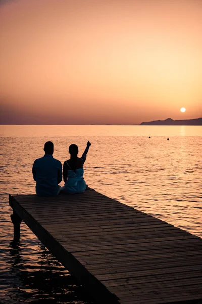 Paar sitzt auf einem Holzsteg und sieht einen bunten Sonnenuntergang am Meer, Männer und Frauen beobachten den Sonnenuntergang auf Kreta Griechenland — Stockfoto
