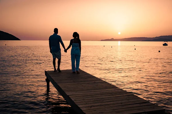 couple seated on a wooden jetty, looking a colorful sunset on the sea , men and woman watching sunset in Crete Greece