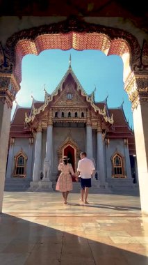 Bir çift, karmaşık mimari ve canlı renklerle çevrili çarpıcı bir Tayland tapınağında romantik bir yürüyüş yapmayı seviyor. Bangkok 'taki Benchamabophit tapınağında güneş parlıyor.
