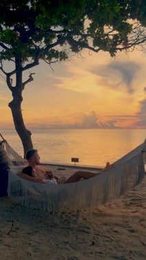 Bir çift, sakin denizi yansıtan altın günbatımının olduğu hamakta huzurlu bir öğleden sonranın tadını çıkarıyor. Sakin atmosfer ve tropikal çevre Tayland 'da mükemmel bir kaçış sağlar..