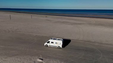 Lakolk Strand, Romo, Danimarka 'da huzurlu bir günde kumlu sahil boyunca ilerleyen, açık mavi gökyüzünün altında mükemmel bir kaçış atmosferi yaratan büyüleyici bir karavan görülüyor..