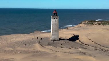 Rubjerknudde deniz fenerinin kıyısına doğru hafif dalgalar süzülüyor, tarihi bir dönüm noktası olan geniş kumlu kumullarla çevrili. Doğa bu manzarayı kucaklarken parlak gökyüzü güzel gölgeler bırakıyor..