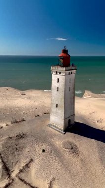 Rubjerg Knude deniz fenerinin büyüleyici görüntüsü Danimarka 'nın kumlu kıyı şeridinde dimdik duruyor. Kuzey Denizi 'nin berrak mavi suları bu tarihi yapı için sakin bir zemin oluşturur..