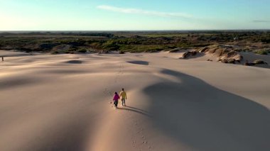Güneş batarken Danimarka 'nın Rabjerg Mile kentindeki kum tepelerinde el ele yürüyen iki insan, sakin manzaranın üzerine sıcak bir parıltı saçıyor ve uzun gölgeler yaratıyor..