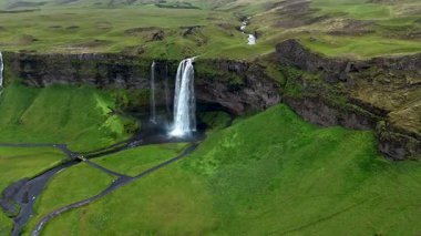Seljalandsfoss şelalesini keşfedin İzlanda 'nın canlı yeşilliklerinde yuva yapmış. Su kayalıkların üzerinden dramatik bir şekilde akarak gün ışığında büyüleyici bir doğa harikası yaratır..