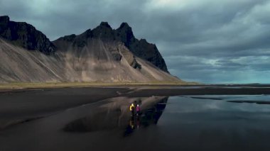 Gün batımında nefes kesici Stoksnes Vestrahorn 'u keşfedin, görkemli dağların sakin sularla buluştuğu yeri. İki maceraperest gökyüzünü yansıtan kara kumlar üzerinde dururken doğanın güzelliğine tanık olun..
