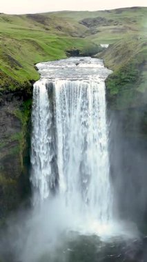 Skogafoss şelaleleri, İzlanda 'daki kayalık bir uçurumdan aşağı, canlı yeşil tepeler ve sisle çevrili. Bu çarpıcı yerde, alacakaranlıkta güçlü görüntüler var..