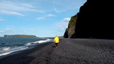 Bir gezgin, İzlanda 'daki Reynisfjara Sahili' nin büyüleyici siyah kumları boyunca yürür. Yükselen kayalıklar ve sakin dalgalar bu sakin sahil kaçışı için nefes kesici bir zemin oluşturur..