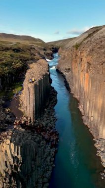 İzlanda 'daki Studlagil Kanyonu' nun nefes kesici güzelliğini deneyimleyin. Yüksek bazalt sütunları ve berrak gökyüzünün altından akan parlak mavi bir nehir..