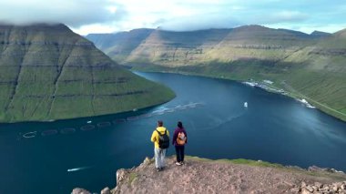 İki yürüyüşçü Klakkur Faroe Adaları 'nda fiyort ve görkemli dağları seyrederek nefes kesici bir manzaranın tadını çıkarıyorlar. Sakin sular gökyüzünü yansıtırken etraflarındaki yemyeşil tepeler dramatik bir şekilde yükseliyor..