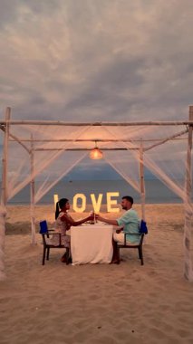 Çiftler Tayland 'da güzel bir kumsal yemeğinde samimi bir anı paylaşırlar. Sıcak günbatımı romantik atmosferi güçlendirir, arka planda büyük bir aşk işareti ile..