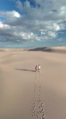 Danimarka 'daki Rabjerg Mil' in büyüleyici kumlu manzarasını keşfedin. İki figür el ele yürür, bulutlarla dolu parlak bir gökyüzünün altında yumuşak kumullarda ayak izleri bırakır..