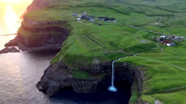 Faroe Adaları 'ndaki Gasadalur köyündeki okyanusa dökülen Mulafossur şelalesinin güzelliğine tanık olun. Altın gün batımı yemyeşil manzarayı ve sakin suları vurgular..
