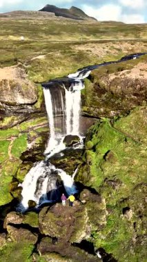 İzlanda 'daki Grundarfoss Şelalesi' nin çarpıcı güzelliğini keşfedin. Bereketli yeşillikler ve kayalık arazilerle çevrili iki figür, çağlayan suyun altında doğal güçten dehşete kapılır..