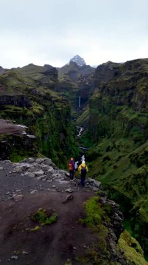 Maceracılar İzlanda 'daki Mulagljufur Kanyonu' nun kenarında durmuş yemyeşil kayalıklara ve uzak şelaleye hayret ediyorlar. Dramatik manzara doğal güzellikleri ve huzuru gözler önüne seriyor..