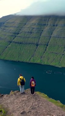 Klakkur 'da, Faroe Adaları' ndaki engebeli bir uçurumun tepesinde, dramatik manzaraya hayran iki yürüyüşçü duruyor. Aşağıda, derin mavi sular parlak yeşil tepelerle tezat oluşturarak çarpıcı bir doğal zemin oluşturuyor..