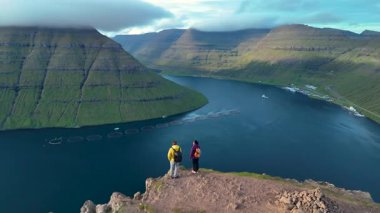Zirveden nefes kesici manzara, iki yürüyüşçünün Klakkur Faroe Adaları 'ndaki yemyeşil uçurumlara ve sakin körfeze baktığını gösteriyor. Manzara, açık bir günde güzelliği sergiliyor..