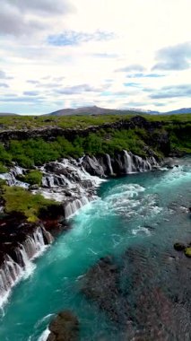 Hraunfossar şelalesinin büyüleyici akışını deneyimleyin, berrak turkuaz sular karanlık lav kayaları üzerinde çağlıyor. Bu doğal mucize İzlandalıların nefes kesici manzaralarını ve zengin biyolojik çeşitliliğini sergiliyor..