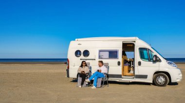 Bir çift, Danimarka 'nın Blokhus Strand kentindeki altın saat boyunca, uzun otlarla çevrili yumuşak kumullarda bir karavanın önünde oturup Kuzey Denizi' nin sakin dalgalarını seyrediyor.